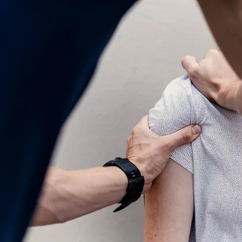 Ein Therapeut umfasst mit beiden Händen die Schulter einer sitzenden Patientin.