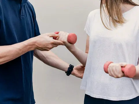 Ein Therapeut korrigiert die Armhaltung einer Patientin bei einer Übung mit leichten Hanteln.